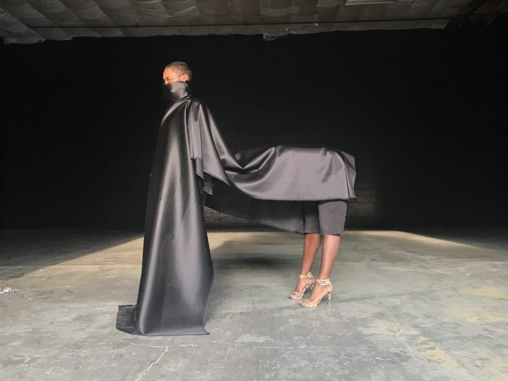 performance still of Dead Thoroughbred (sidony o'neal and keyon gaskin). The performers form the rough shape of a horse, draped in silky black fabric. one performer, as the "legs" of the horse, wears high heels, the other performer, as the "head" of the horse, has the bottom half of their face covered with the same silky fabric.