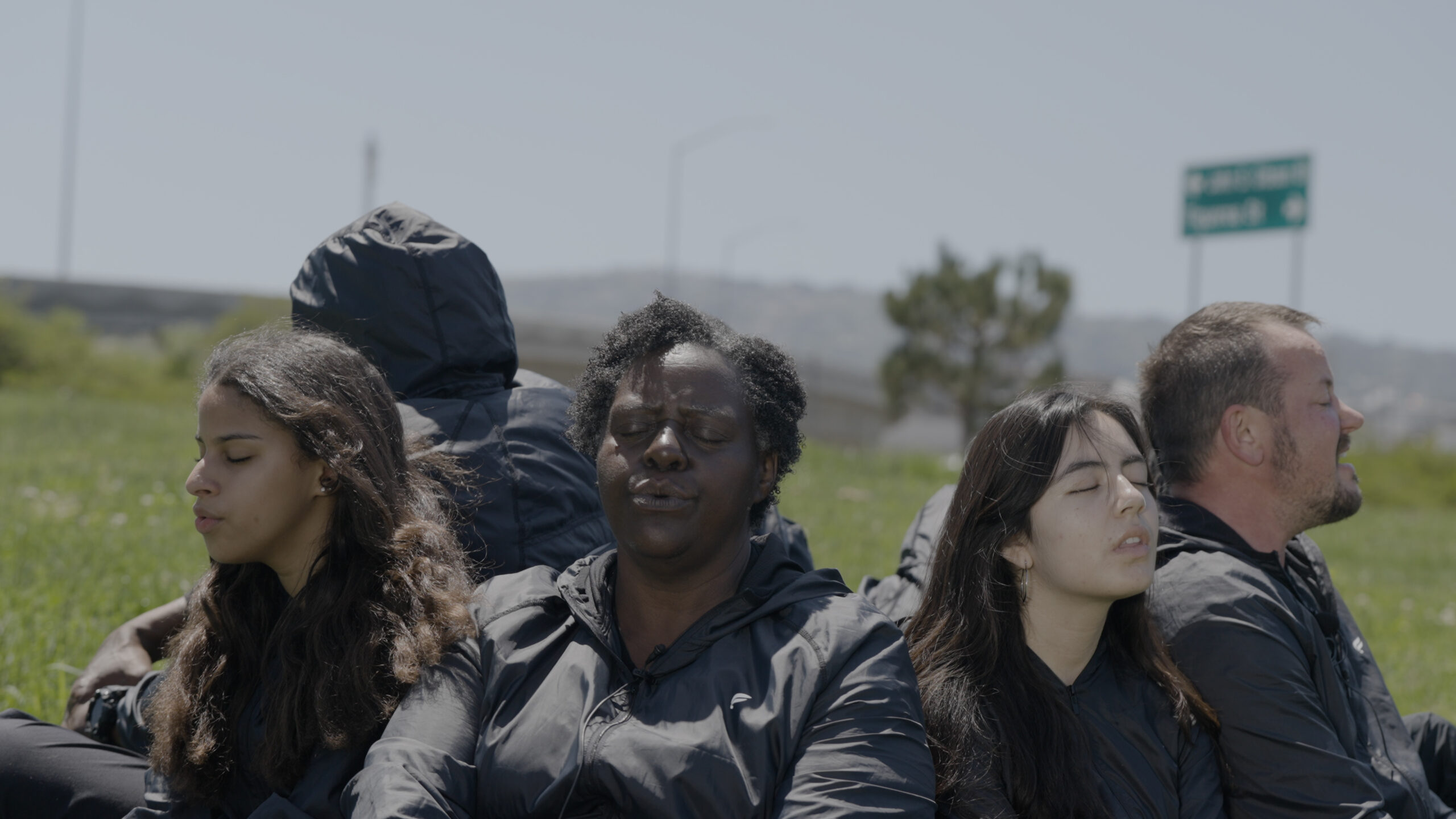 A group of people sit in a circle, facing outward. They appear to be engaged in breathwork or singing, and sittings in grass.