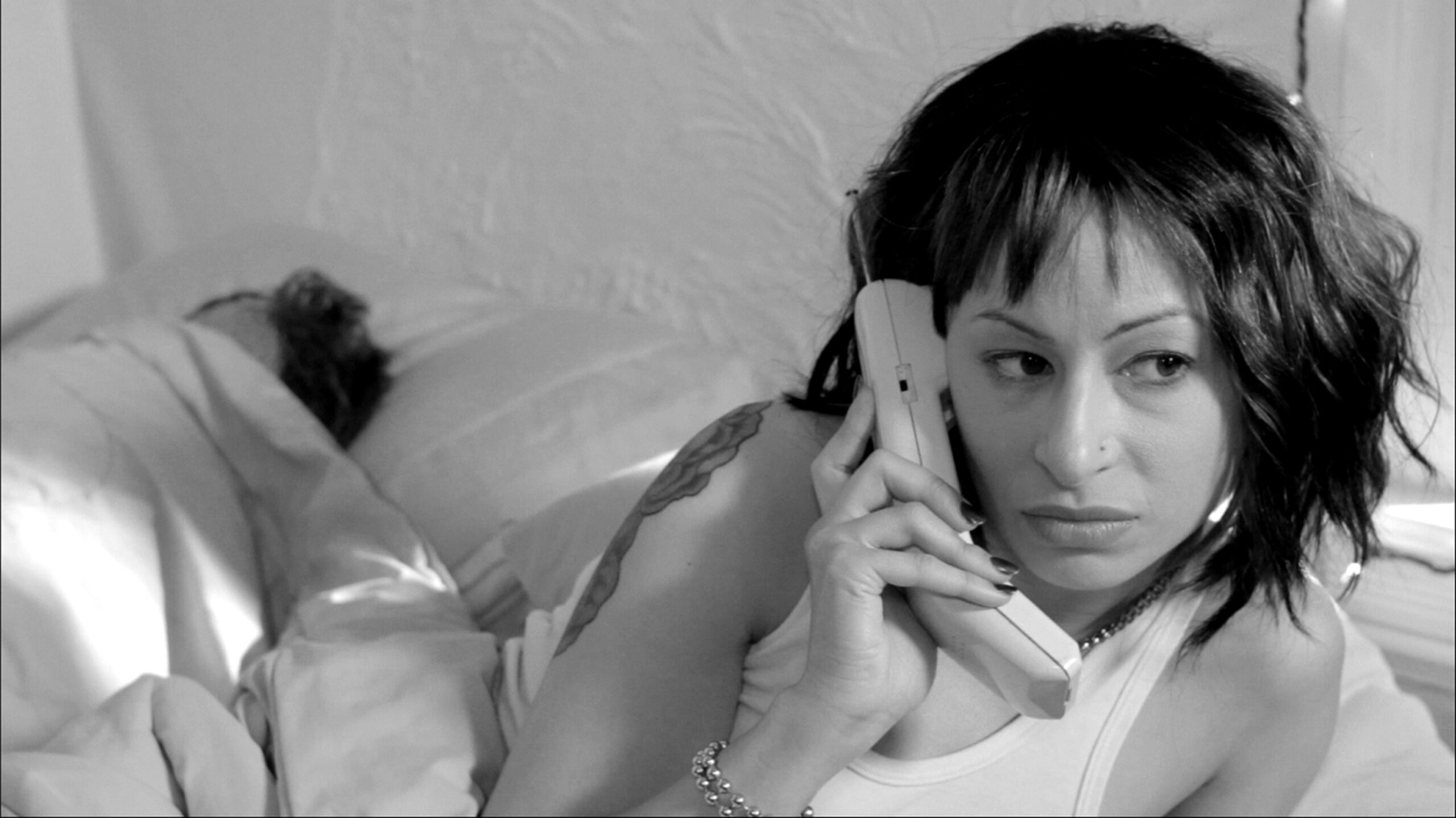 Black and white photo of woman is speaking on a phone. She is in bed. In the background we see that someone else is in the bed sleeping.