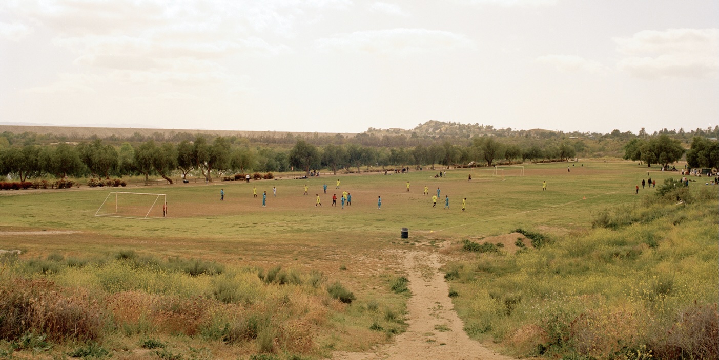A photograph which shows a dirt path leading to the field in the distance. There is a game being played there.